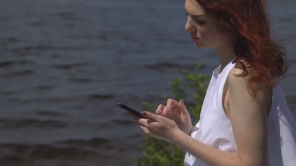 Primer plano de hermosa chica pelirroja en un vestido de verano blanco utiliza un teléfono celular sentado en la orilla y los ríos — Vídeos de Stock