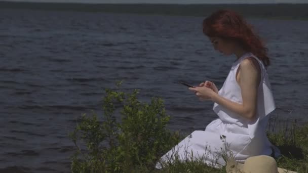 美しい赤い髪の少女の白い夏ドレスは海岸や河川上のソーシャル ネットワークで座って携帯電話を使用してのクローズ アップ — ストック動画