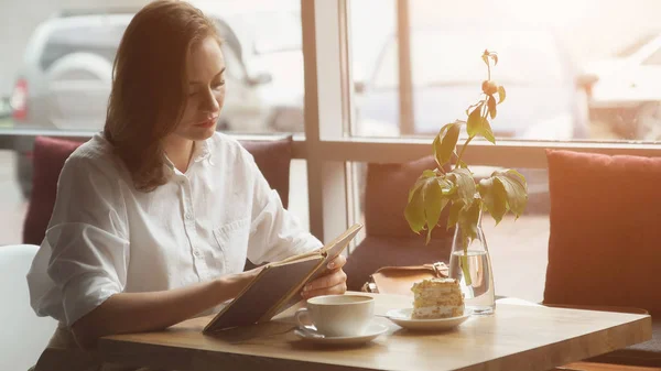 여자는 카페에 앉아 커피와 케이크를 즐기고 책 읽으십시오. — 스톡 사진