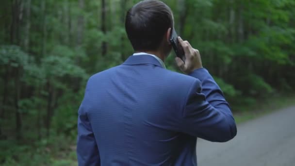 Красивый мужчина идет по дороге в лесу, разговаривает по телефону, смотрит назад — стоковое видео