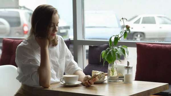 Una chica usa un teléfono celular sentado en un café. una mujer joven envía un mensaje con teléfono móvil y beber café —  Fotos de Stock
