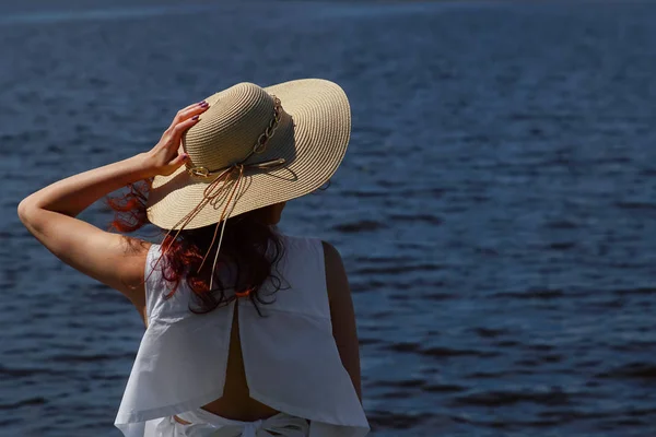 Pessoa feminina atraente em vestido branco segurando chapéu de glamour ao ar livre na margem do rio, visão traseira, água azul profunda no fundo. copypace — Fotografia de Stock