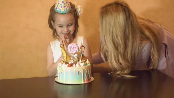 Madre e hija soplan velas en un pastel de cumpleaños. el cumpleaños de una niña. la cumpleañera en el sombrero de fiesta en la velada infantil —  Fotos de Stock