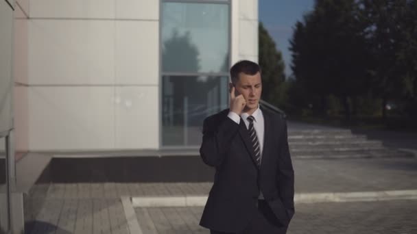 Attraktiver Geschäftsmann im schwarzen Anzug, der vor dem Hintergrund eines Business Centers im Freien mit dem Smartphone spricht. — Stockvideo