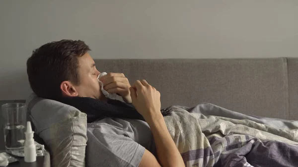 man lying sick in bed next to his medications in his home and blowing his nose in a handkerchief