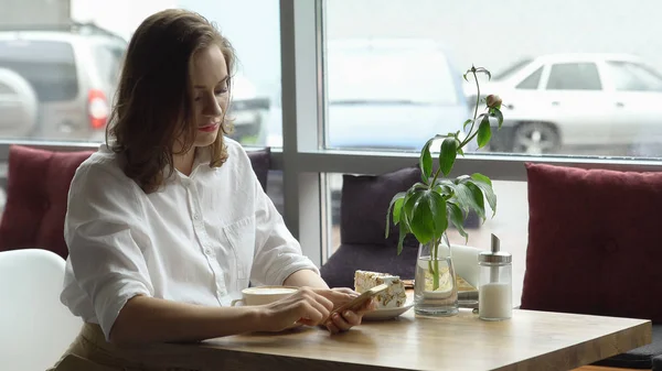 Una chica usa un teléfono celular sentado en un café. una mujer joven envía un mensaje con teléfono móvil y beber café —  Fotos de Stock