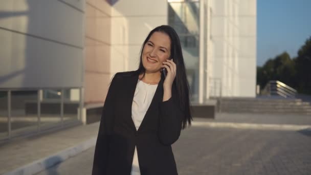 Joven mujer de negocios en traje negro hablando en el teléfono inteligente contra el telón de fondo del centro de oficina en el día soleado — Vídeos de Stock