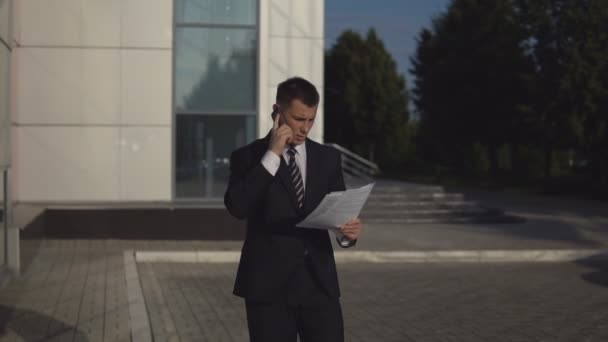 Бизнесмен в черном костюме разговаривает на смартфоне и выбрасывает документы на задний план здания — стоковое видео