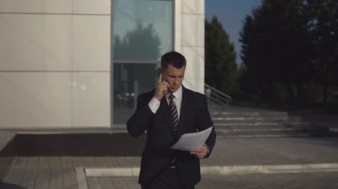 Genç işadamı smartphone üzerinde konuşmak ve Vesaik mukabili güneşli günde arka bina iş dışarı atmak