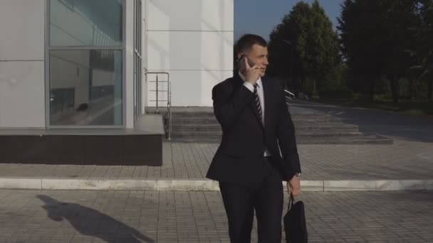 Серьезный красивый бизнесмен в черном костюме с дипломатом разговаривает по смартфону и спешит на встречу на фоне бизнес-центра под открытым небом — стоковое видео