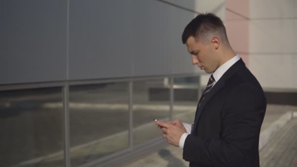 Young businessman typing text on smartphone standing on background of business building outdoor. — Stock Video