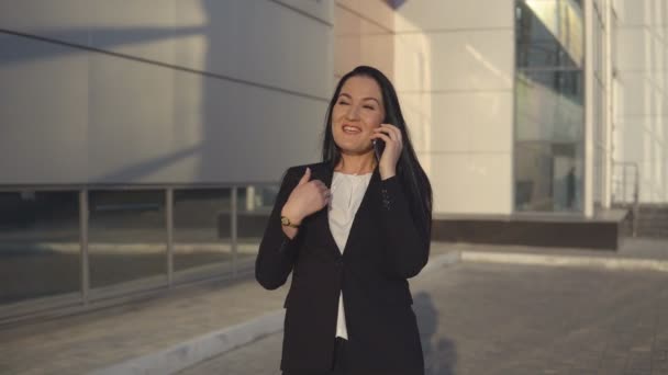 Chica joven en traje negro hablando en el teléfono inteligente contra el telón de fondo del centro de oficina al aire libre — Vídeos de Stock