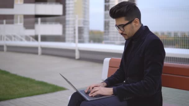 Atractivo hombre de negocios en traje negro con portátil sentado al aire libre — Vídeos de Stock