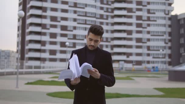 Atractivo joven hombre de negocios en traje negro tirar documentos al aire libre contra el edificio de oficinas . — Vídeos de Stock
