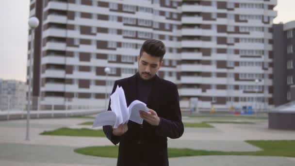 Atractivo joven hombre de negocios en traje negro tirar documentos al aire libre contra el edificio de oficinas . — Vídeo de stock