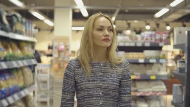 Chica joven está caminando cerca de la estantería del supermercado y selecciona los productos — Vídeos de Stock