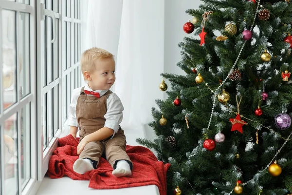 Der kleine Junge sitzt am Fenster und schaut auf den Weihnachtsbaum. Urlaub, Geschenk und Neujahrskonzept — Stockfoto