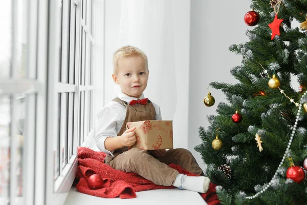 Kleiner Junge mit Geschenk in der Hand sitzt am Weihnachtsbaum und blickt in die Kamera. — Stockfoto
