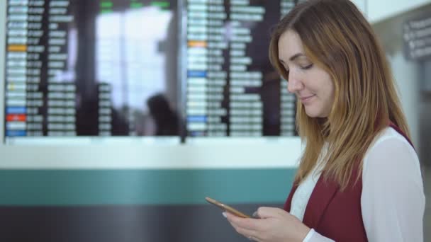 Красива дівчина стоїть на тлі розмитого табло з повідомленням і вибором польоту в смартфоні . — стокове відео
