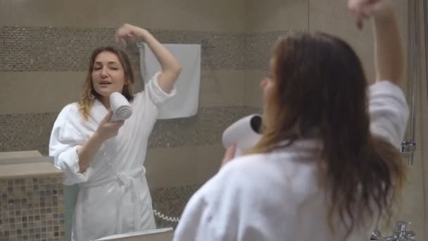 Vicces bájos lány fehér köntöst vett fürdő és tükör hajszárítóval előtt énekel. — Stock videók