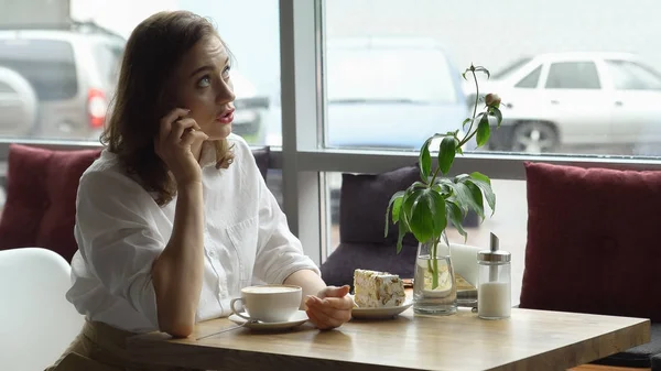 Una chica usa un teléfono celular sentado en un café. una mujer joven envía un mensaje con teléfono móvil y beber café —  Fotos de Stock