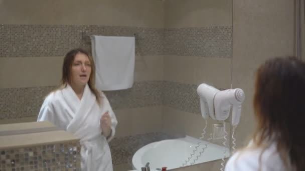 Grappig charmante meisje in witte robe nam bad en zingt voor spiegel met wc en haardroger. — Stockvideo