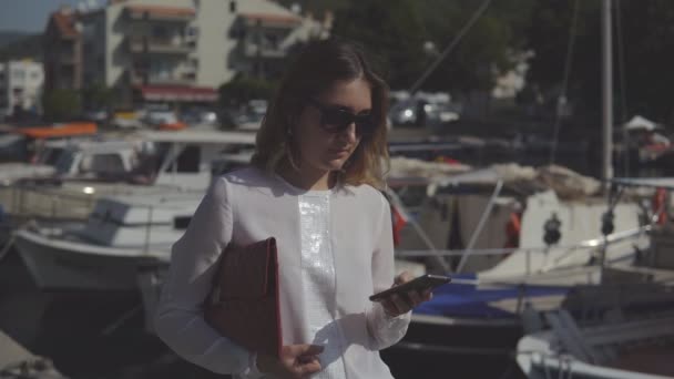 Positive Geschäftsfrau mit Smartphone und Handtasche in formeller Kleidung, die auf dem Hintergrund verschwommener Boote steht. — Stockvideo