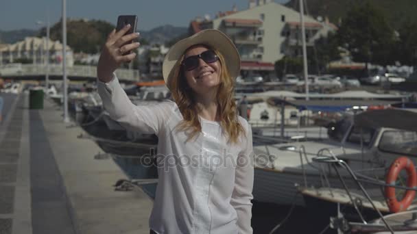 Mooie jonge meisje in hoed en zonnebril maakt selfie staande op de pier op zonnige zomerdag. — Stockvideo