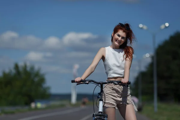 Рыжая молодая девушка в шортах и топах позирует стоя с велосипедом — стоковое фото