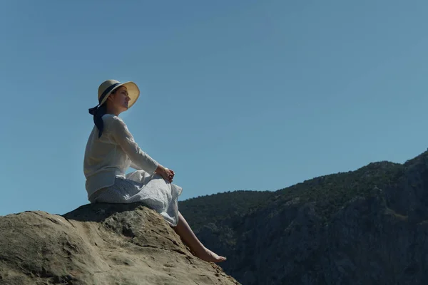 Junges Mädchen mit Hut sitzt auf einem hohen Steinhügel und genießt die schöne Natur vor dem Hintergrund der Hügel an einem sonnigen Sommertag. — Stockfoto