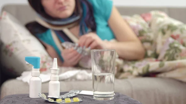 Tablets, spray and water on the chair and young sick woman takes a pill in bed on the background. — Stock Photo, Image