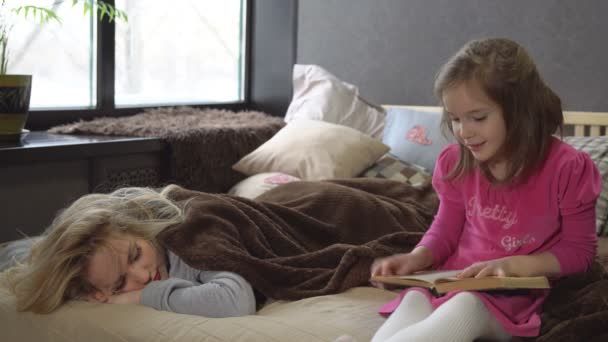 Дочь читает книгу на кровати, пока мама спит. — стоковое видео