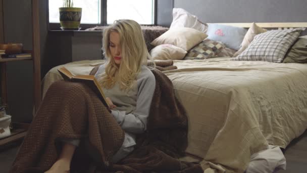 Joven rubia está sentada en el suelo en una manta junto a la cama y está leyendo un libro — Vídeos de Stock