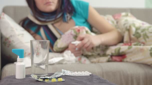Таблетки, спрей и вода на стул и молодая больная девушка принимает лекарство от соплей на заднем плане . — стоковое видео