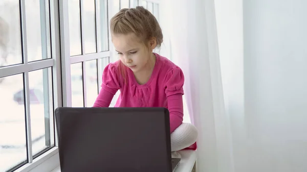 Klein meisje in een roze jurk zit bij het raam met een laptop. — Stockfoto
