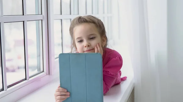 Pembe elbiseli küçük kız bir tablet ile cam kenarında oturan — Stok fotoğraf