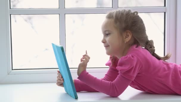 핑크 드레스에 어린 소녀는 태블릿 창이 앉아 있다. 측면 보기 — 비디오