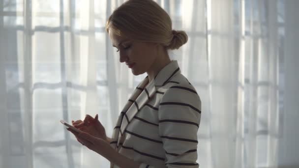 Молодая женщина в полосатой куртке с телефоном на фоне окна — стоковое видео