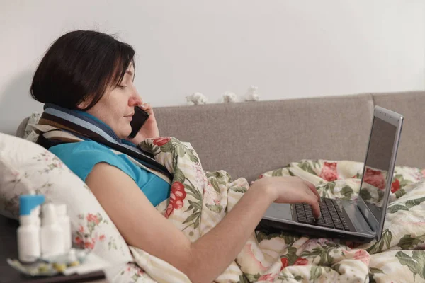 Mujer enferma acostada en la cama en casa, hablando por teléfono y trabajando en el portátil —  Fotos de Stock