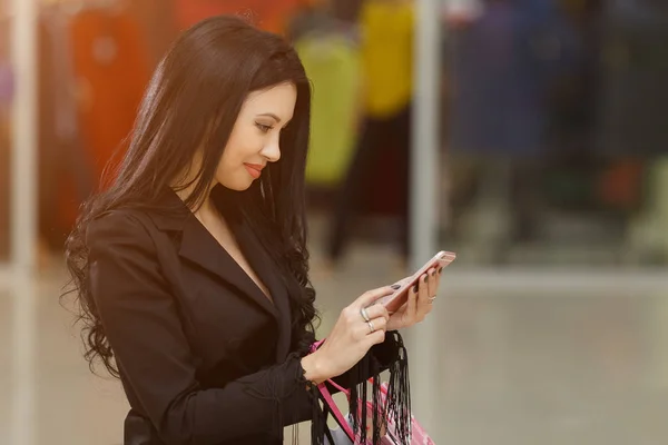 Csinos, fiatal lány gazdaság bevásárló táskák, és használ egy okos telefon, Mall. — Stock Fotó