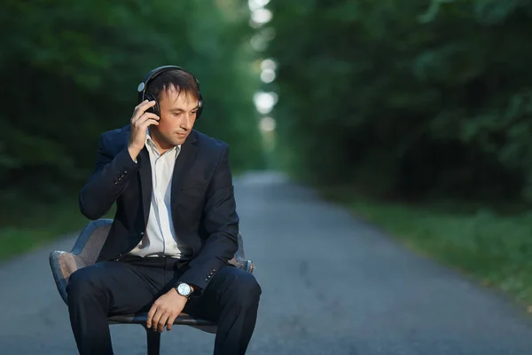 Joven hombre de negocios sentado en una silla en un traje escuchando música en los auriculares en medio del bosque — Foto de Stock