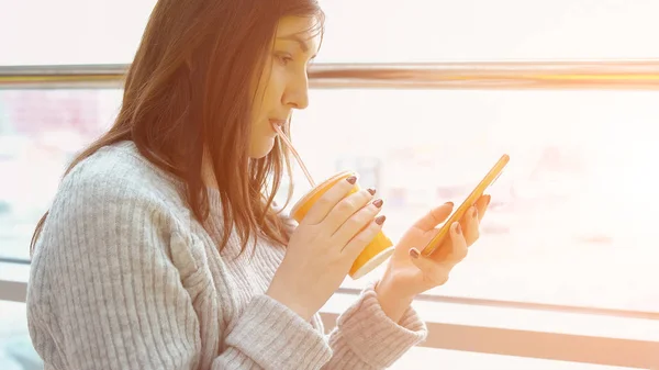 Fiatal nő egy szürke pulóver egy telefon, egy kávézóban, az ablakon, ivás lé — Stock Fotó