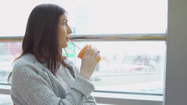 Mujer joven bebe jugo recién exprimido en un café, mirando por la ventana — Vídeo de stock