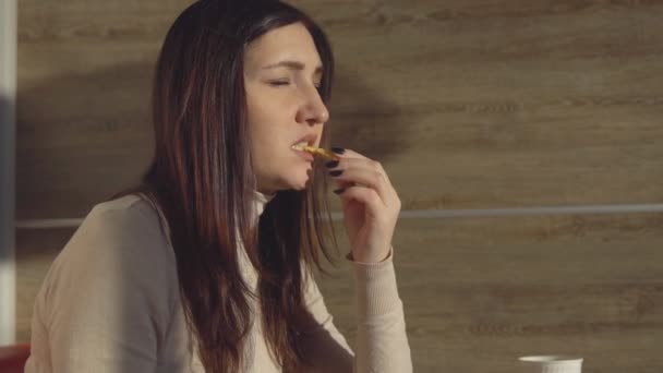 Fiatal barna nő kávézóban eszik sült krumpli. A koncepció az egészségtelen ételek — Stock videók