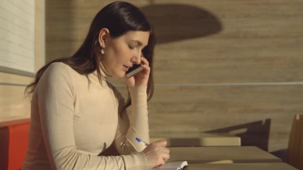 Giovane donna in un caffè che parla al telefono e prende appunti in un quaderno — Video Stock