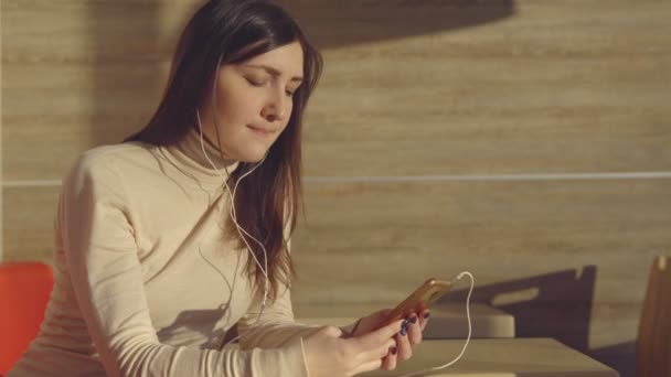 Mujer joven sentada en la cafetería con auriculares y escuchando música — Vídeos de Stock