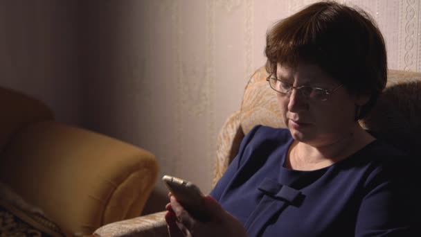 Vrouw in een blauwe jurk en bril zit in een fauteuil met een telefoon — Stockvideo