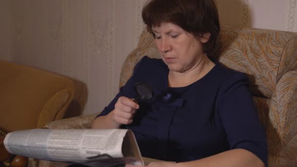 Middle-aged woman reads a newspaper through a magnifying glass sitting in a chair — Stock Video