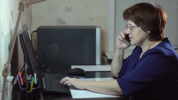 La donna che indossa gli occhiali mentre lavora al computer sta parlando al telefono — Video Stock