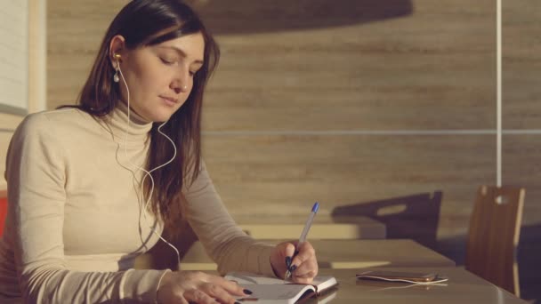 Jonge vrouw zitten in cafe met koptelefoon en het maken van aantekeningen in notitieblok — Stockvideo